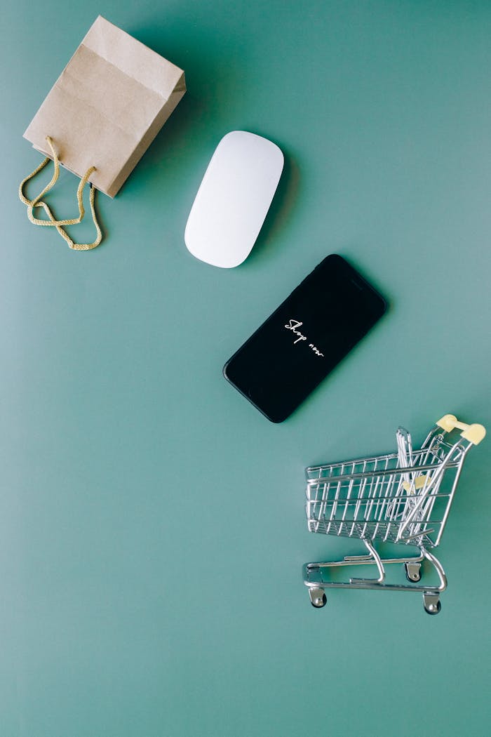 Top view of a smartphone, mouse, paper bag, and mini shopping cart on a green background, symbolizing e-commerce.